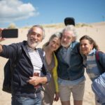 A joyful group of senior friends taking a selfie at a sandy beach in Portugal under clear blue skies.